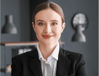 A person in a black blazer and white shirt smiles confidently in an office setting with gray walls and modern lighting.