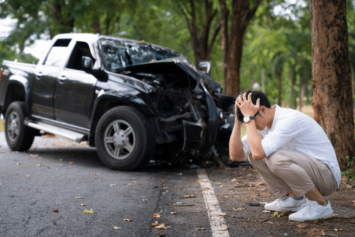 A man crouches with his head in his hands near a black pickup truck with a damaged front, on a tree-lined road.