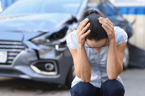 A person crouches with hands on head in front of a damaged car.