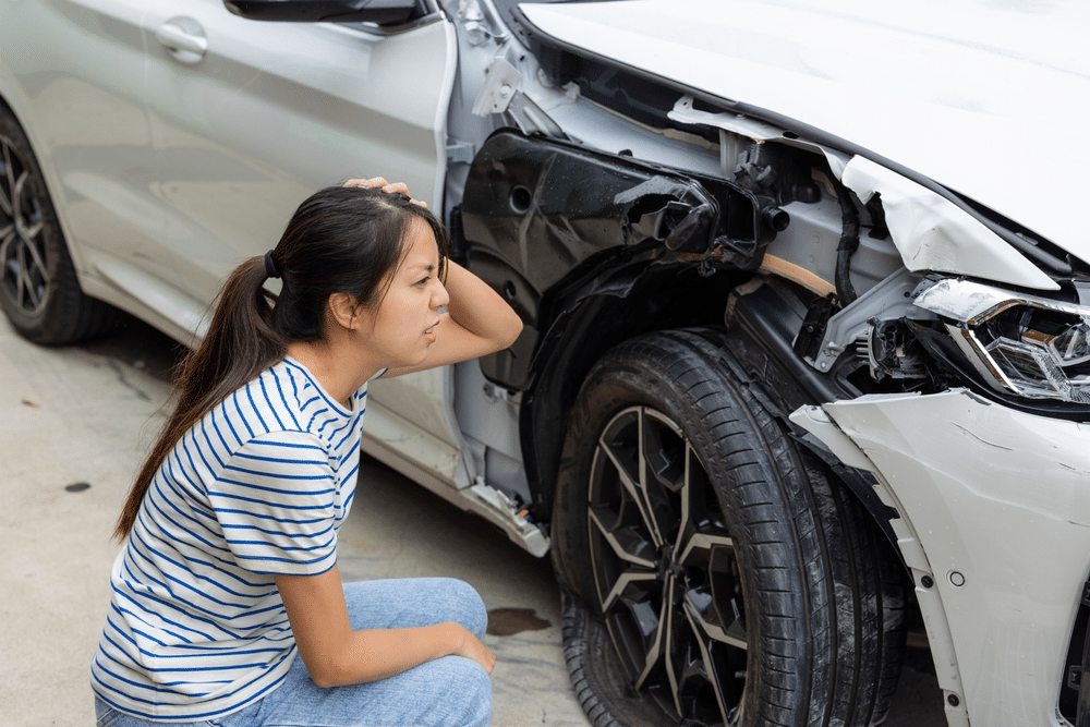 Person examines damage on the front of a white car, including dents and a misaligned wheel, while kneeling and holding their head.