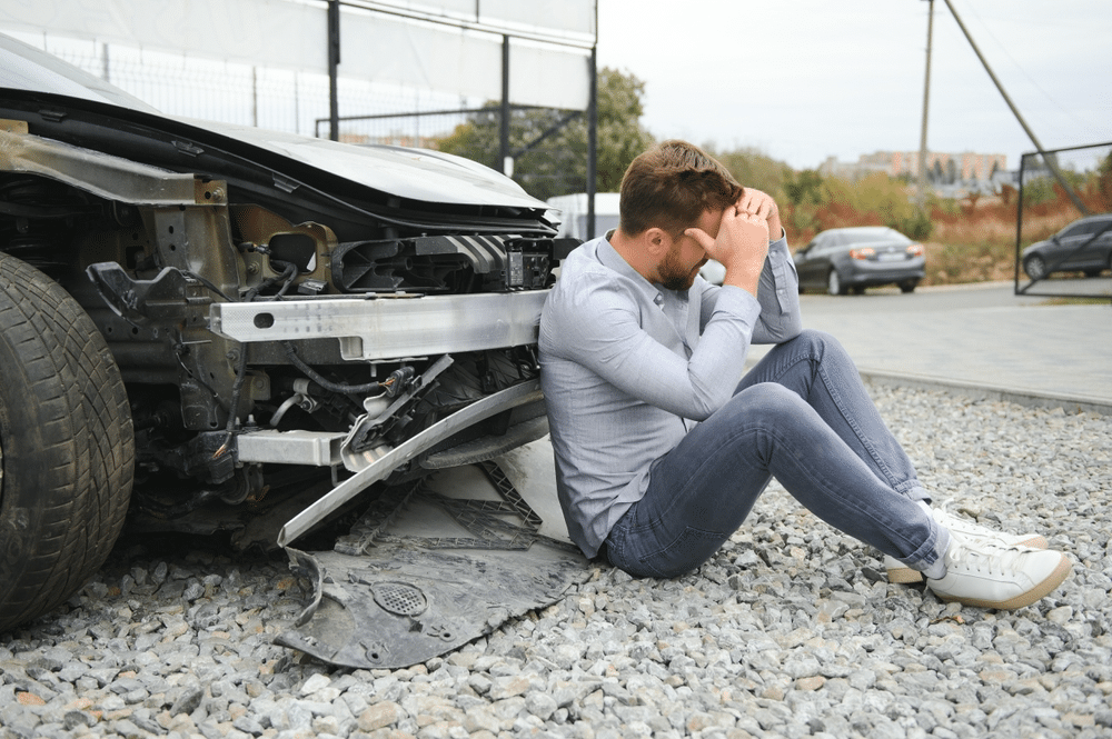 Man sitting on gravel next to a damaged car, holding his head in his hands, appears distressed.