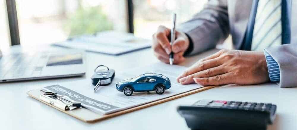 A person in a suit signs a contract at a desk with a toy car, keys, and calculator nearby.