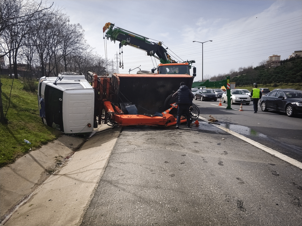 Two overturned vehicles on roadside, attended by a tow truck and several people. Nearby cars are passing by, and traffic cones are placed for safety.