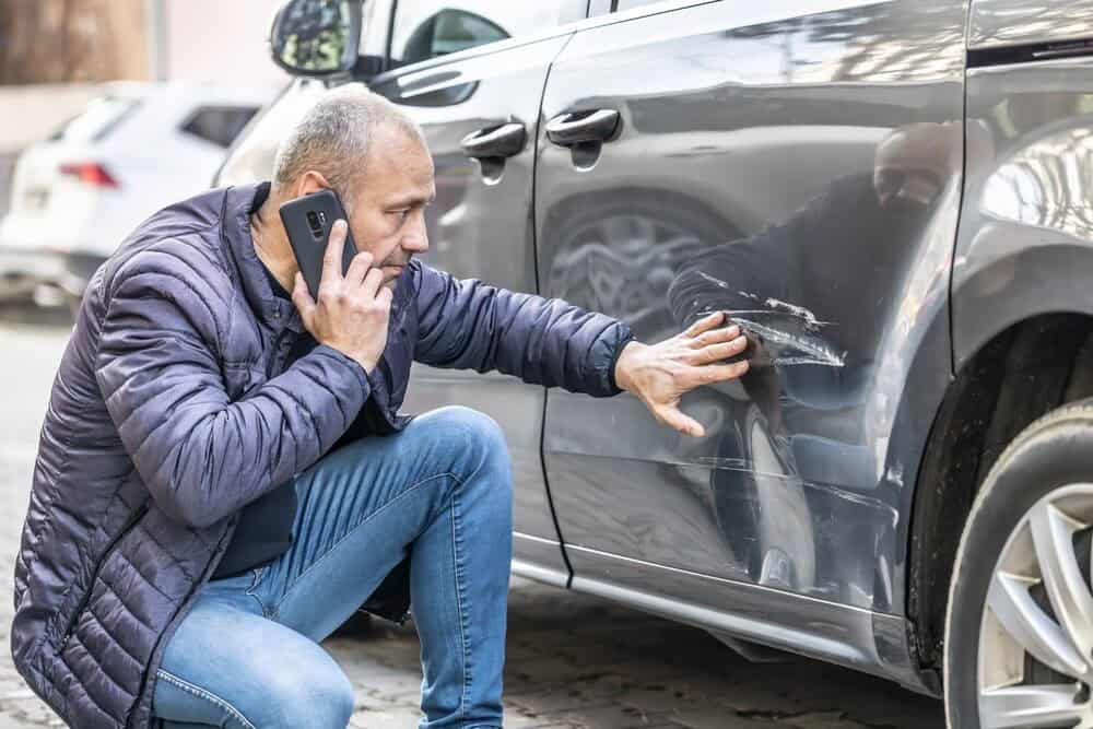 Man inspecting car scratch while talking on the phone, kneeling by a damaged vehicle on a street.