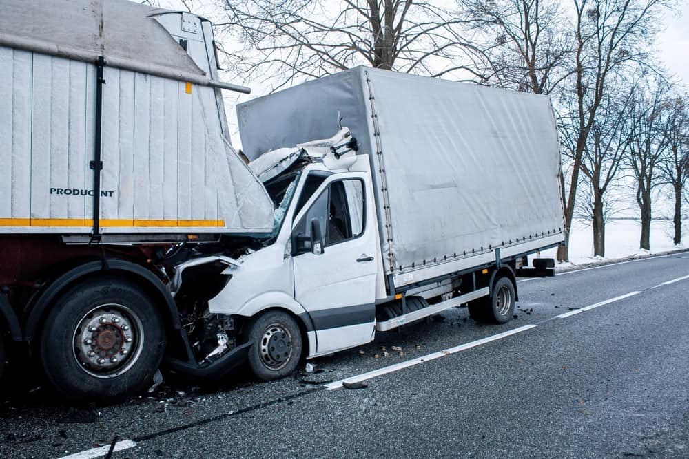A white delivery van is crashed into the back of a larger truck on a snowy road. Trees line the roadside.