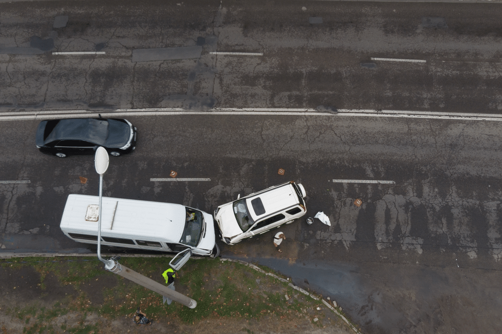 Aerial view of a two-vehicle collision involving a white van and a white SUV on a multi-lane road. A person in a vest is near the scene, and two debris bags are visible on the ground.