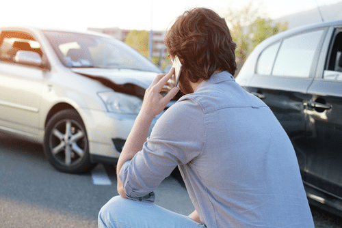 A man on the phone looks at two cars with damaged fronts in a parking lot.
