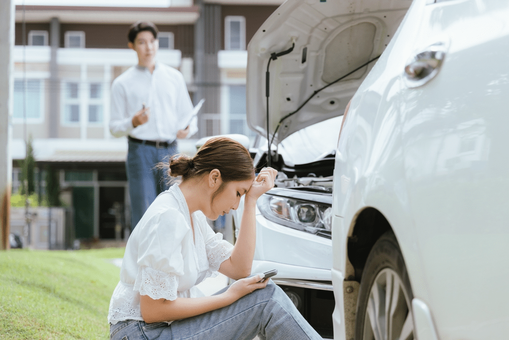 A woman sits on the grass looking at her phone beside a car with an open hood. A man stands in the background, holding papers.