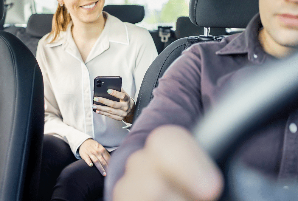 A person in the back seat of a car smiles while looking at their phone. The driver is focused on the road.
