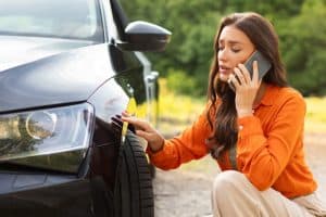 A woman in an orange blouse kneels beside a car, talking on a cellphone, examining the car's front bumper. Trees are visible in the background.