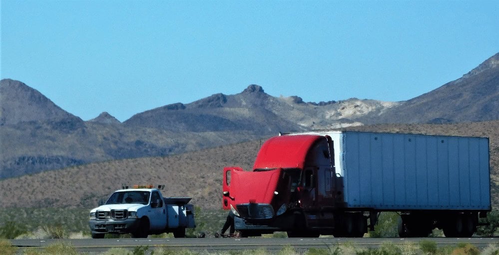 A red semi-truck with an open hood is parked on a desert road next to a white utility truck. Mountains are in the background.